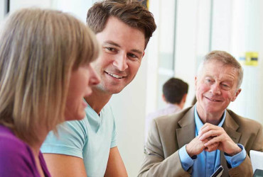 A young man smiles while his mother discusses her hearing aid experience to her audiologist